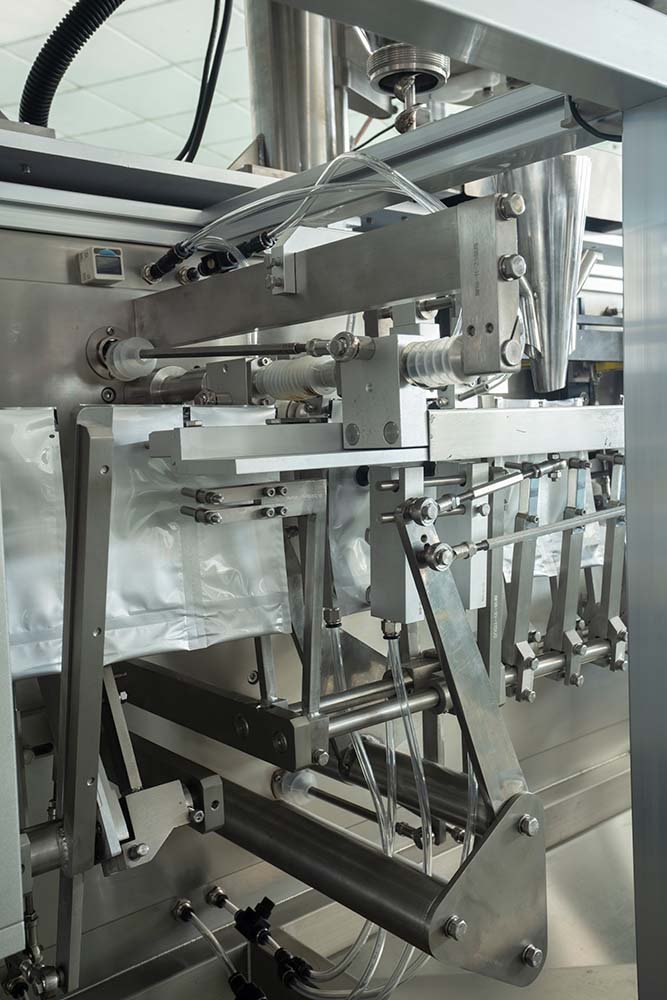 Technical detail of the INMAYPACK HS Series doypack shaping system, showing pneumatic mechanical levers stretching and opening the bottom gusset of stand-up laminate pouches before the filling nozzle descends.