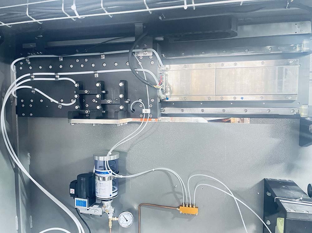 Internal compartments of a packaging machine showing orderly pneumatic tubings, an electric lubrication pump with a pressure gauge, and a linear motor drive track.
